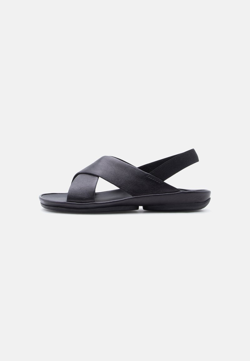 Sandale en cuir noir avec des brides croisées et une bride élastique à l'arrière, semelle plate et rembourrée, vue latérale sur fond blanc uni.