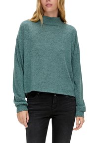 Turkusowy krótki sweatshirt z wysokim kołnierzem, miękkim teksturowanym materiałem i opadającymi ramionami, w połączeniu z ciemnymi dżinsami.