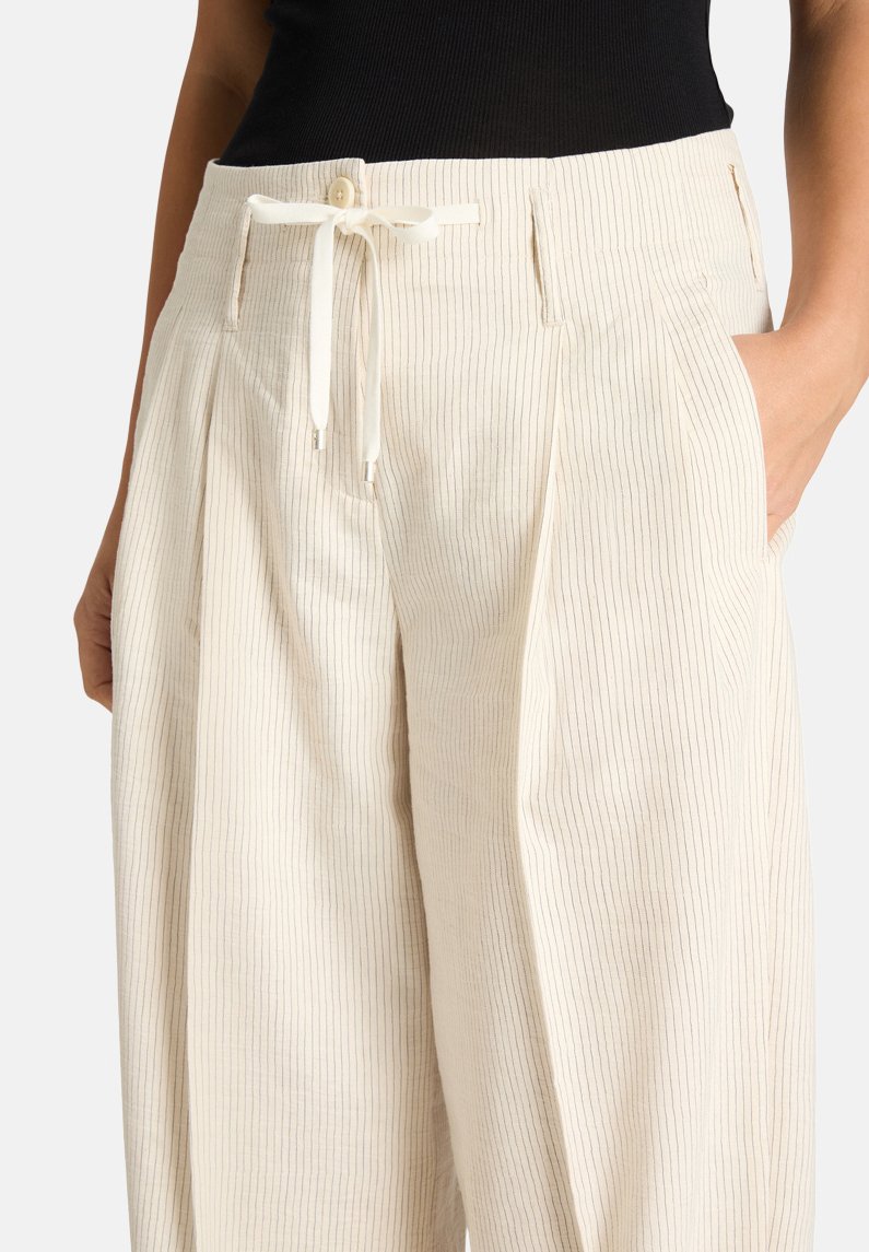 Frau trägt cremefarbene, weit geschnittene Hose mit dünnen vertikalen Streifen, einem taillierten Bund mit Knöpfen, einem Kordelzug und Seitentaschen, in denen sie die Hände hat.