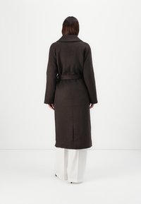 Long brown coat made of textured fabric, featuring a belted waist, wide sleeves, and a notched collar. Back view shows a vent at the hem.