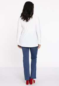 Weißes Langarmoberteil mit abgerundetem Saum, kombiniert mit blauen Bootcut-Jeans und roten High Heels. Einfaches Design, glatte Textur.