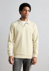 Bézs hosszú ujjú póló V-nyakú kivitelben. Puha anyag, laza szabás és bordázott mandzsetták. Fehér alapréteg fölött viselve.