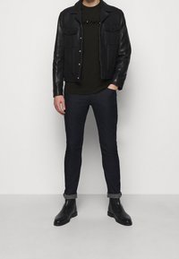 Uomo che indossa una giacca di pelle nera, una t-shirt con logo nera, jeans scuri arrotolati e stivali di pelle neri, in piedi contro uno sfondo bianco.