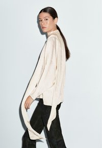 Maglietta beige chiaro a maniche lunghe con vestibilità ampia e maniche fluide, abbinata a pantaloni neri. Tessuto dalla texture liscia, con dettagli minimi.