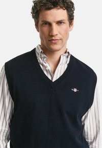 Uomo con capelli ricci che indossa un gilet di maglia blu scuro con scollo a V sopra una camicia bianca con sottili strisce verticali rosse.