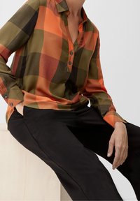 Camicia a quadri arancione e verde con colletto, dotata di chiusura con bottoni e maniche lunghe, abbinata a pantaloni neri, seduta su una superficie chiara.
