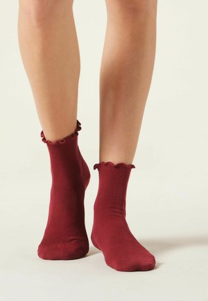 Burgundyröda bomullssockor med ribbad textur och vågiga, scallopade toppar, visade på nakna ben mot en ljus bakgrund.