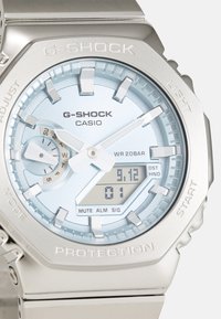 Silver G-SHOCK watch with a light blue dial, digital display, and stainless steel band. Features include subdial, prominent markers, and protective casing.