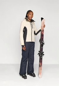 Beige és fekete sídzsekit, valamint fekete nadrágot viselő nő állva tart egy pár fekete kötésekkel ellátott sílécet.