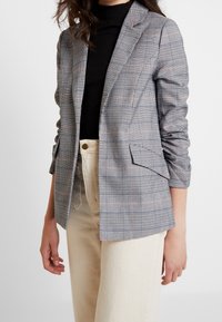 Blazer à carreaux gris et blanc avec des accents orange subtils ; doté d'un col cranté, de deux poches avant et d'une coupe ajustée.