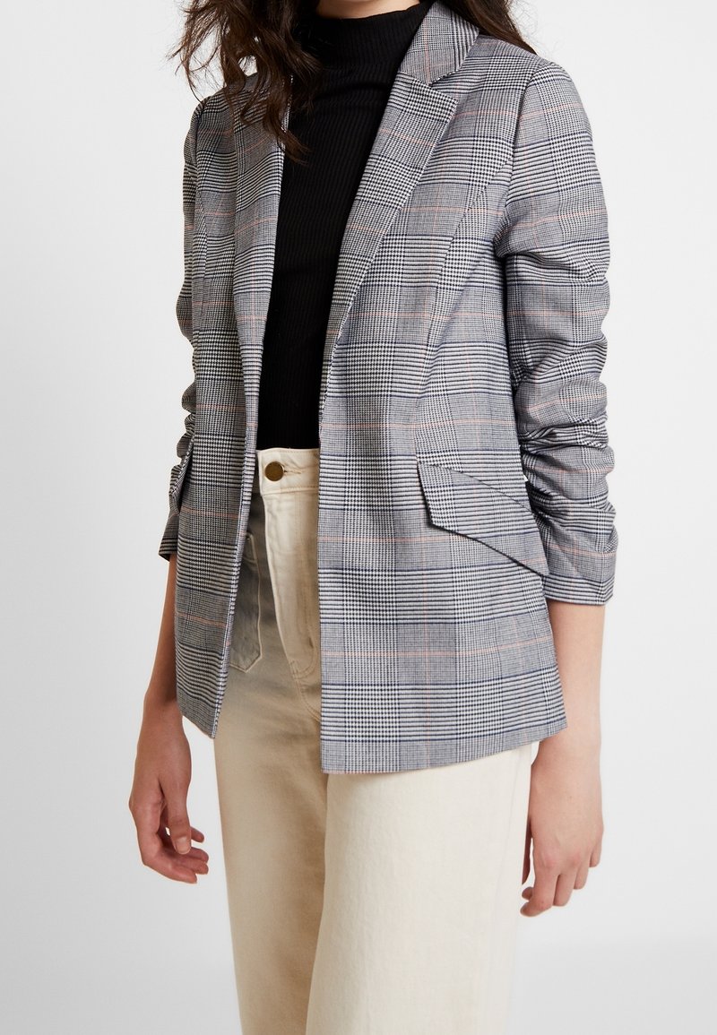 Blazer à carreaux gris et blanc avec des accents orange subtils ; doté d'un col cranté, de deux poches avant et d'une coupe ajustée.