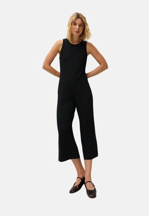 Mujer de pie con las manos detrás de la espalda, vestida con un mono negro sin mangas y zapatos Mary Jane negros sobre un fondo claro y liso.