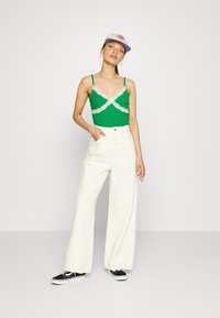 Top verde com rendas, calças brancas de perna larga, ténis pretos e um chapéu branco com detalhes vermelhos. Modelo posa com uma mão no bolso.