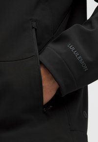 Svart Lululemon-jacka med en slät textur, slim fit, ficka med dragkedja och subtil logotyp på ärmen.