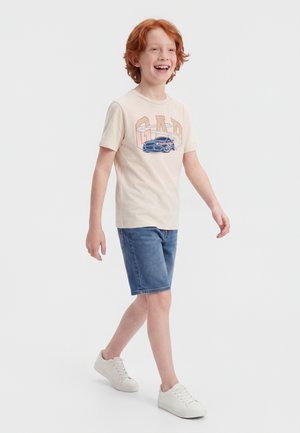Băiat cu părul roșcat mergând, purtând un tricou bej cu grafică de mașină, pantaloni scurți din denim albastru și adidași albi, zâmbind pe fundal alb.