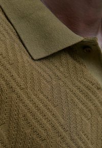 Närbild av en brun texturerad stickad polo-tröja med krage och knapp på en persons hals och övre bröst.