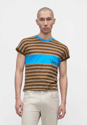 Jeune homme aux cheveux courts portant un t-shirt rayé marron avec une bande horizontale bleue et un pantalon beige clair sur fond blanc.