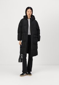 Musta lasitakki huppulla, etuvetoketjulla, kahdella sivutaskulla ja pehmeällä pinnalla. Päällä valkoinen paita ja mustat housut.