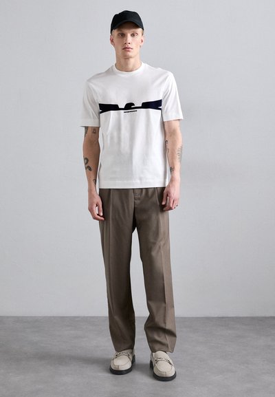 Witte T-shirt met korte mouwen en marineblauwe accenten, beige broek en lichtgekleurde schoenen. Eenvoudig ontwerp met zichtbare tattoo's op de armen.