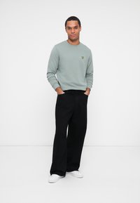 Felpa verde menta con logo giallo, abbinata a pantaloni neri larghi e sneakers bianche. Design semplice e tessuto testurizzato.