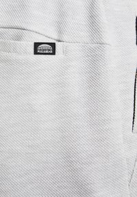 Tissu texturé gris clair avec un motif de tissage subtil, comportant une petite étiquette noire avec "PULL&BEAR" sur une poche.