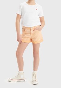 Camiseta blanca de algodón con un pequeño logo rojo, combinada con pantalones cortos de mezclilla color durazno claro con dobladillos enrollados y zapatillas altas beige.