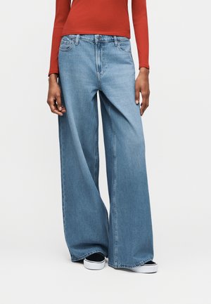 Άτομο που φοράει ψηλόμεσα, ανοιχτό μπλε τζιν με φαρδιά πόδια, μαύρα και λευκά αθλητικά παπούτσια και μακρυμάνικη κόκκινη μπλούζα, στέκεται μπροστά σε απλό φόντο.