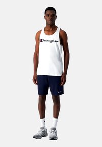 Hvid tanktop med "Champion" logo i marineblå rød skrift, parret med marineblå shorts, grå sneakers og hvide sokker, stående på en neutral baggrund.