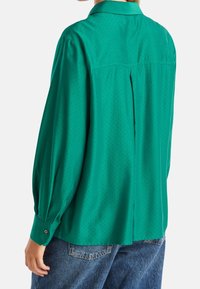 Grön långärmad skjorta med knappmanchett, rundad nederkant och subtil prickig mönstring; tillverkad av lättviktig tyg med en mjuk yta.