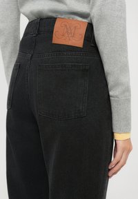 Jeans in denim nero con una finitura testurizzata, dotati di due tasche posteriori e di una patta in pelle marrone con marchio in rilievo sul cinturino.