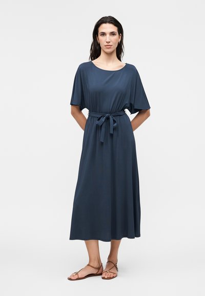 Femme debout portant une robe midi bleu marine avec une ceinture nouée à la taille et des manches courtes, associée à des sandales plates marron sur fond blanc.