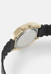 Fekete gumi szíj aranyszínű rozsdamentes acél tokkal és két koronanyomógombbal. Simább hát lapka a márka logójával gravírozva.