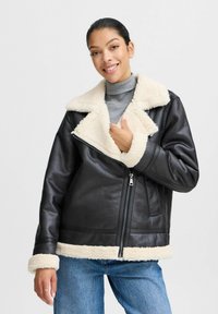 Melnā ādas jaka ar pūkainu beidzīgu šērlina apkakli un aprocēm, ar rāvējslēdzēju un sānu kabatām. Valkāta virs pelēka turtlenecka un ziliem džinsiem.
