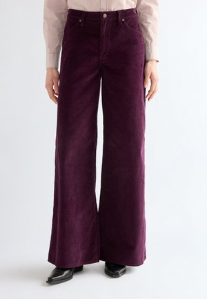 Femme portant un pantalon large en velours côtelé violet foncé et des chaussures noires à bout carré, debout devant un fond clair uni.