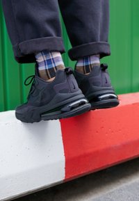 Personne portant des baskets noires et des chaussettes à carreaux se tient sur une barrière en béton peinte en rouge et blanc contre un mur vert.