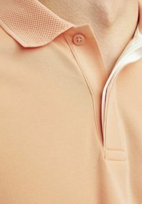 Polo tričko v světlém broskvovém odstínu s texturovaným límečkem. Obsahuje přední knoflík a hladkou texturu látky, která zdůrazňuje detaily kolem výstřihu.