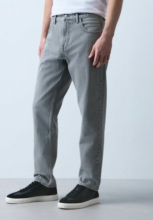 Mand iført lysegrå jeans og sorte sneakers med hvide såler, stående mod en ensfarvet lys baggrund.