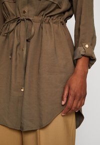 Olivově zelená košile s dlouhým rukávem, elastickým pasem, zlatými knoflíky, límečkem a zaobleným lemem. Ruka položená na boku.