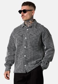 Schwarz-weiß gestreiftes Hemd mit Kragen, Knopfleiste und Brusttasche, kombiniert mit schwarzen Sonnenbrillen und dunkler Hose.