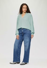 Lichtgroene blouse met lange mouwen en V-hals, gecombineerd met blauwe, wijde jeans en zilveren enkellaarsjes. Smooth stof en een ontspannen pasvorm.
