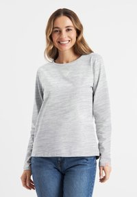 Langärmliges graues Oberteil aus strukturiertem Stoff, mit rundem Ausschnitt, kleinem Herzdetail auf der Brust und seitlichen Schlitzen. Kombiniert mit blauen Jeans.