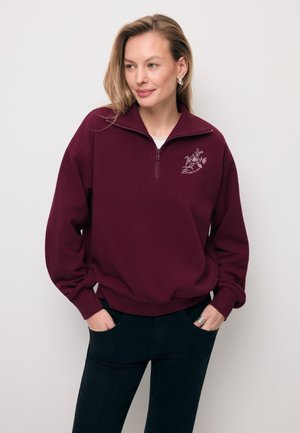 Kobieta o blond włosach, ubrana w bordową bluzę na ćwiartkę z haftem kwiatowym na piersi oraz czarne spodnie, z rękami w kieszeniach.
