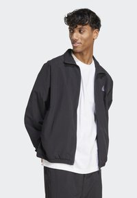 Svart lättvikts zip-up jacka med elastiska ärmslut, kombinerad med en vit t-shirt. Anmärkningsvärt logotypdetalj på vänster bröst. Slät textur.