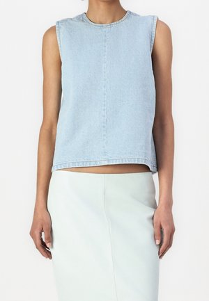 Femme portant un haut en denim bleu clair sans manches associé à une jupe de couleur claire, debout devant un fond blanc uni.