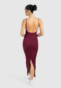 Mujer con cabello rizado recogido en un moño, vestida con un vestido sin mangas, de espalda descubierta y color burdeos, con una abertura en la parte trasera, de espaldas y con zapatillas blancas sobre un fondo liso.