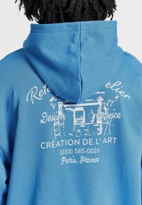 Modrá mikina s kapucí s bílým potištěným textem a designem na zádech, která ukazuje umělecké téma s kontaktními údaji.