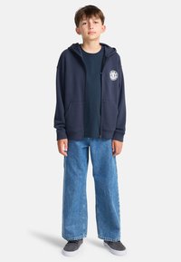 Tmavomodrá mikina na zips s logom vpredu, v kombinácii s tmavomodrým tričkom a svetlomodrými džínsami s širokými nohavicami. Neformálny vzhľad dopĺňa pohodlná obuv.