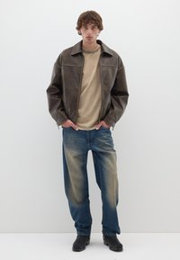 Giacca di pelle marrone con un delicato lucido, t-shirt beige, jeans in denim blu sbiaditi e zoccoli neri, indossati in un ambiente neutro.