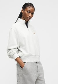 Νεαρή γυναίκα με πλεξούδες στα μαλλιά, φοράει λευκό φούτερ με φερμουάρ στο ένα τέταρτο και ριγέ παντελόνι, στέκεται με το ένα χέρι στην τσέπη.