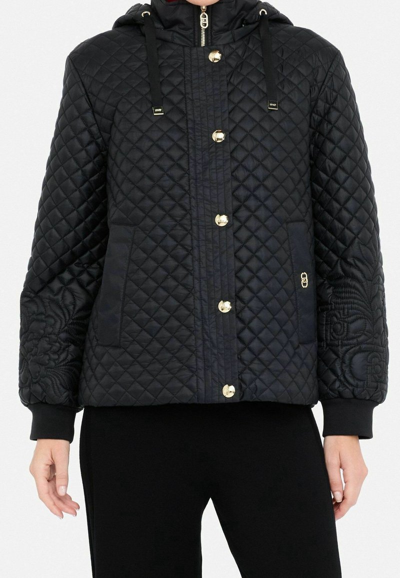 Chaqueta negra acolchada con capucha, botones dorados, dos bolsillos laterales y un bordado floral en el lado izquierdo. Puños acanalados.